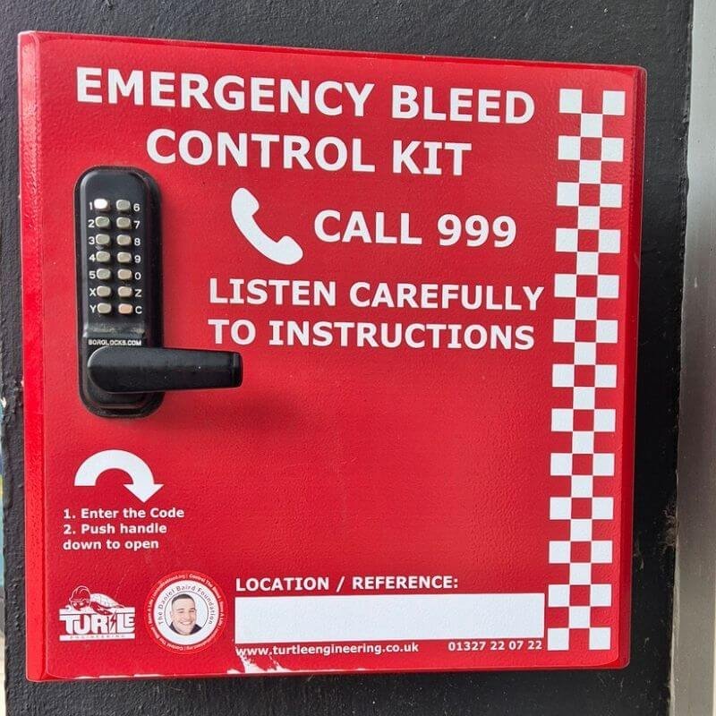Red emergency bleed-control kit box with a keypad lock; bold white text reads 'Call 999' and 'Listen carefully to instructions'